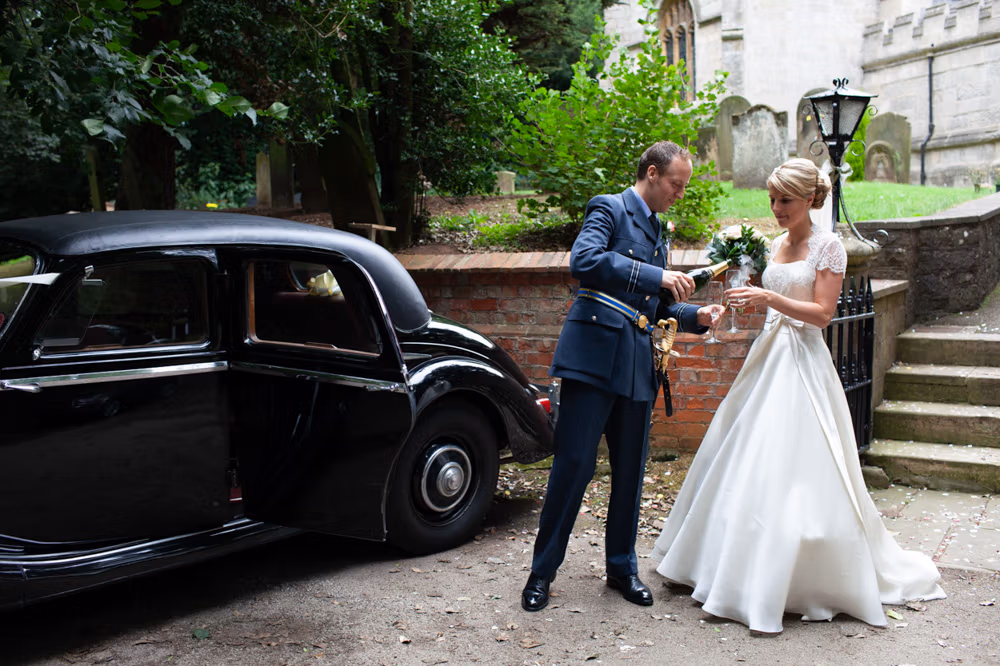 Natalie and Richard - a calm candid moment - alone together as guets have left for the venue - the groom pours champagne for the bride - aside the vintage black wedding car.
