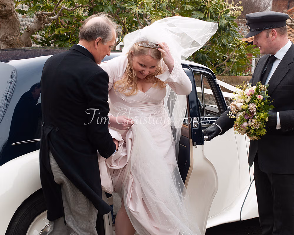 Bride stepping from a vintage car as wind lifts her veil, smiling on arrival.