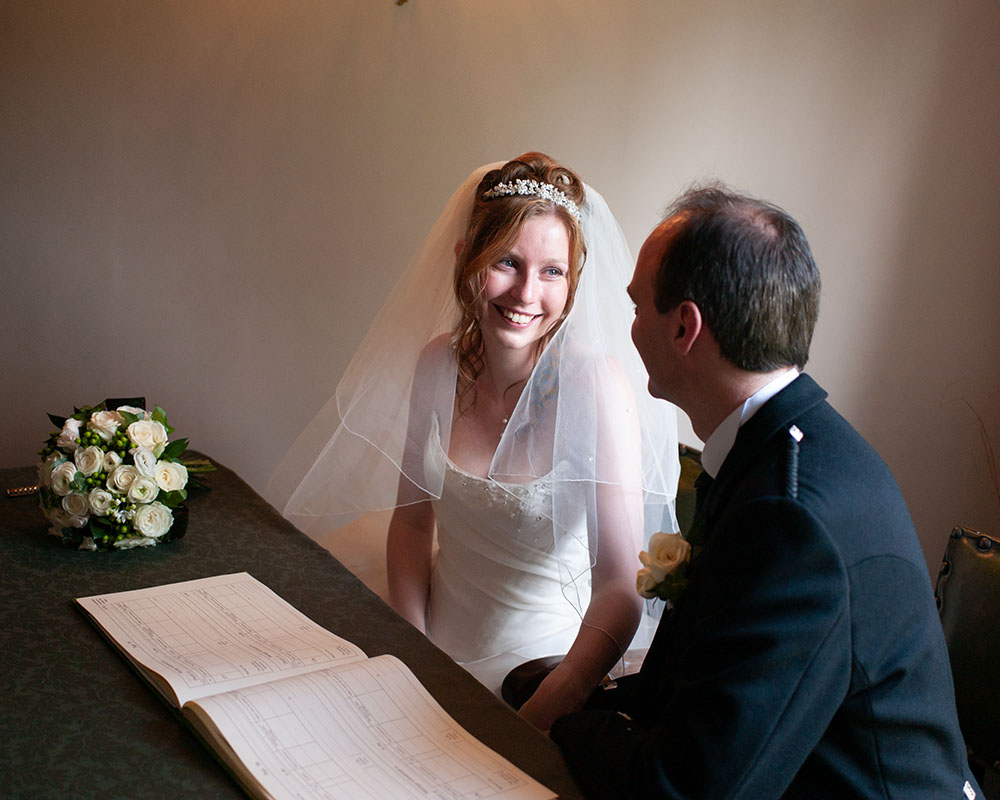The couple sit side by side at the wedding register table, smiling at one another in beautiful natural light, with a rose bouquet nearby.