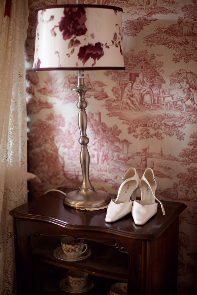 Bridal shoes on a vintage cabinet beside a lamp in the Soughton Hall bridal suite, lace curtains behind