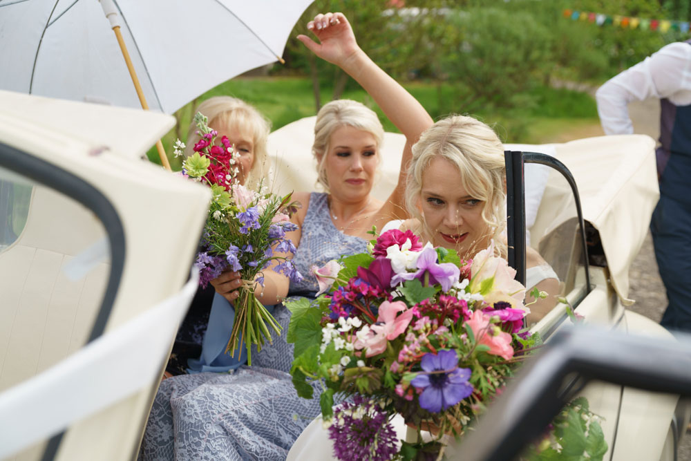 The bride and her bridesmaids step from a vintage cream wedding car, holding soft summer bouquets and a white umbrella — just in case.