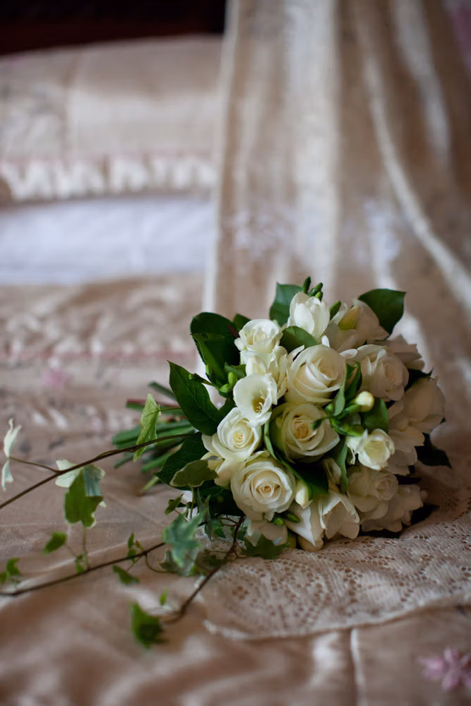 Cream rose bridal bouquet with soft greenery placed on a lace-draped four-poster bed