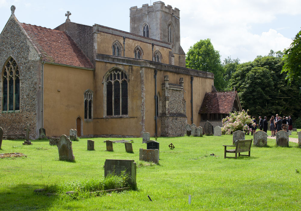 An old pastel-plastered church stands softly in the landscape, pale green grass and empty benches waiting quietly before the ceremony.