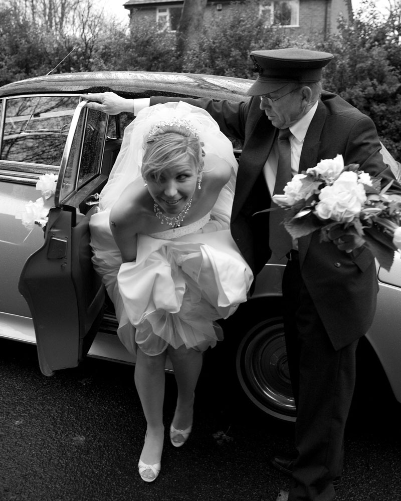 The bride lifts her dress and steps quickly from a vintage car, escaping the rain with a light grasp of the fabric and purpose in her stride.
