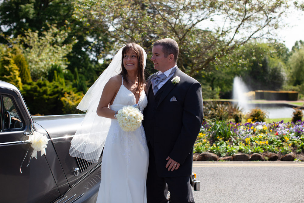 Couple pause by a vintage car near green trees, flowers and fountain in soft colours.