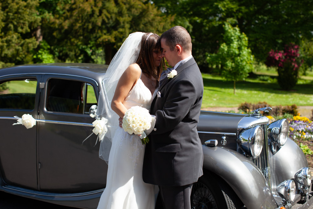 Bride and groom nose to nose by the wedding car in soft green summer tones.