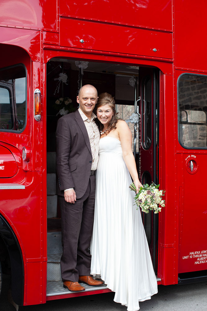 Couple smile widely on the steps of their red double decker wedding bus.