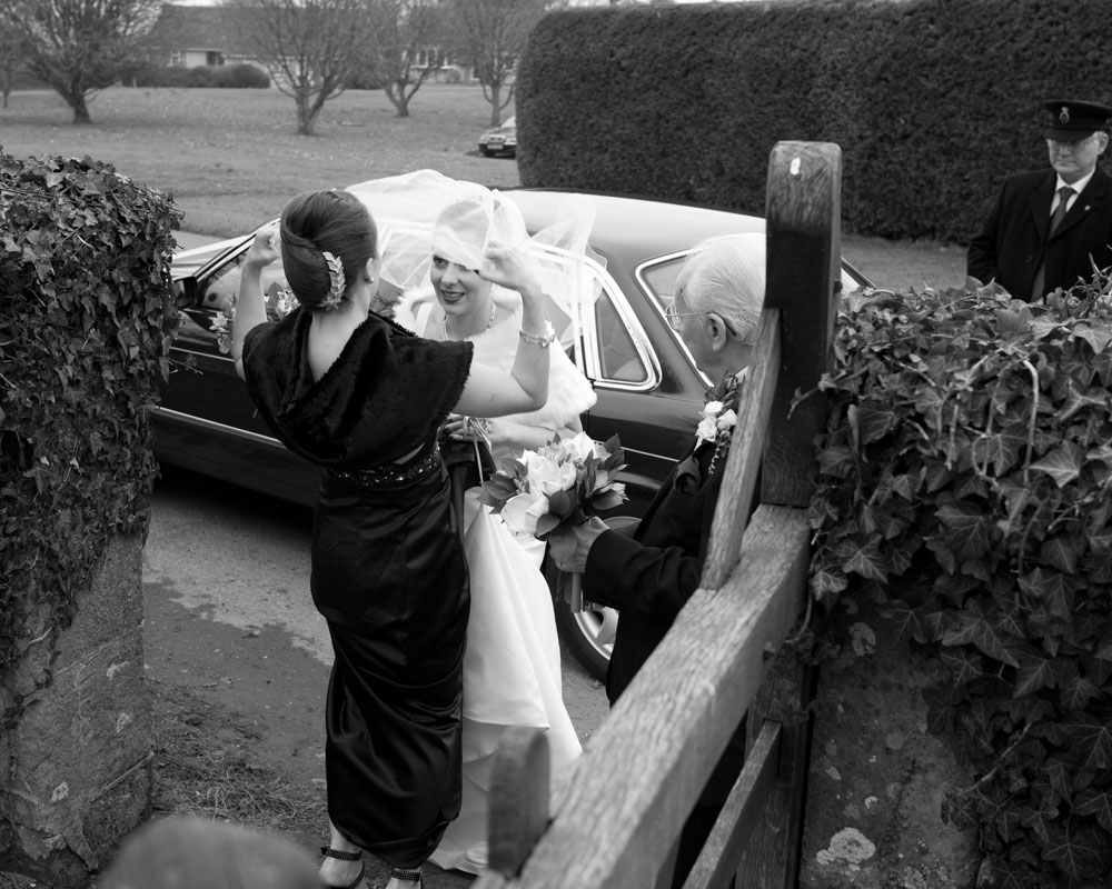 In black and white, the bride’s sister adjusts her veil at the gate of a small church as their grandfather quietly watches from behind.