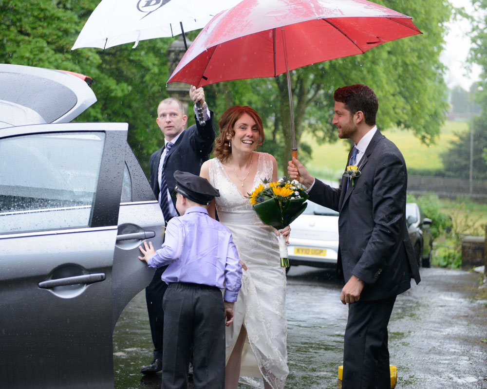 The bride smiles softly, her expression turned upward as she stands in the rain — a quiet moment of joy just after arrival.
