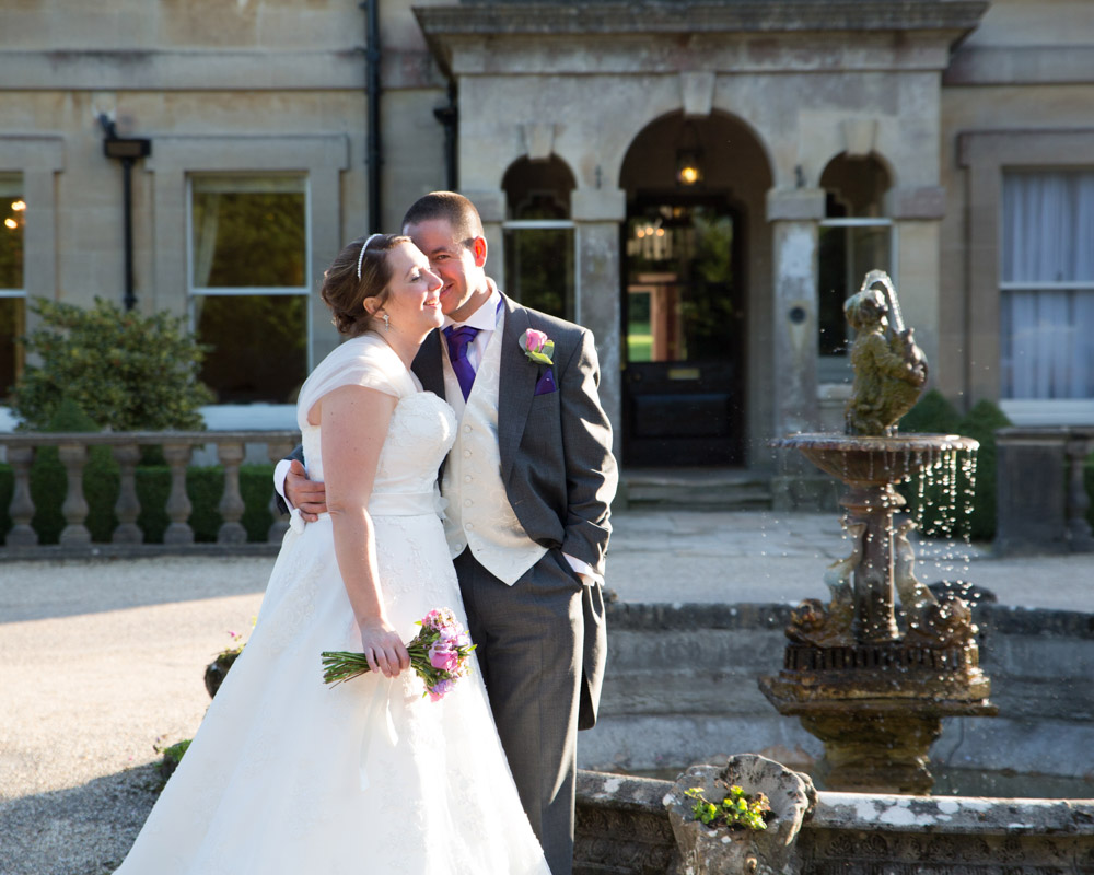 Groom smiles nervously in October sunshine near a water feature at a historic venue.