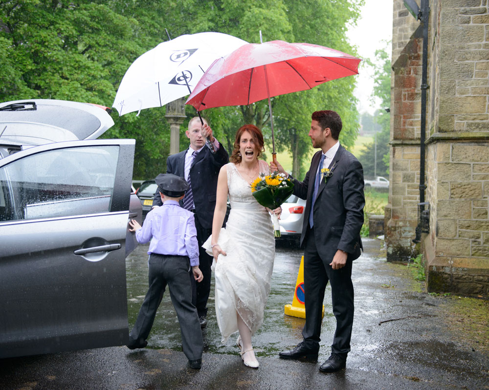 The bride arrives and lets out a wide-eyed gasp at the falling rain, while groomsmen hold umbrellas around her.