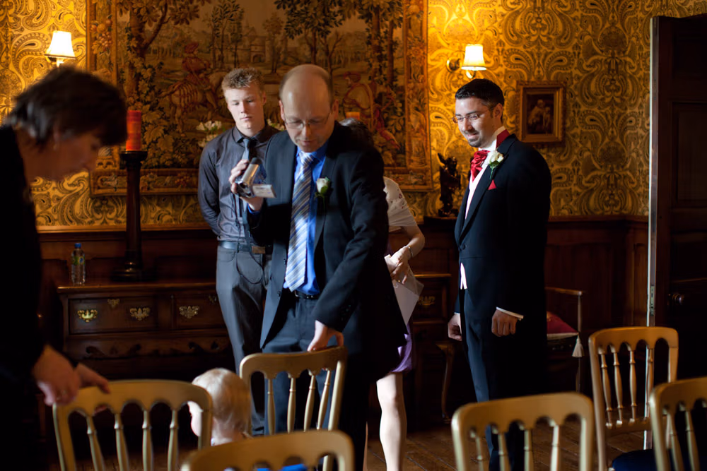 Registrar preparing with video camera filming, guests chatting, groom smiling nearby