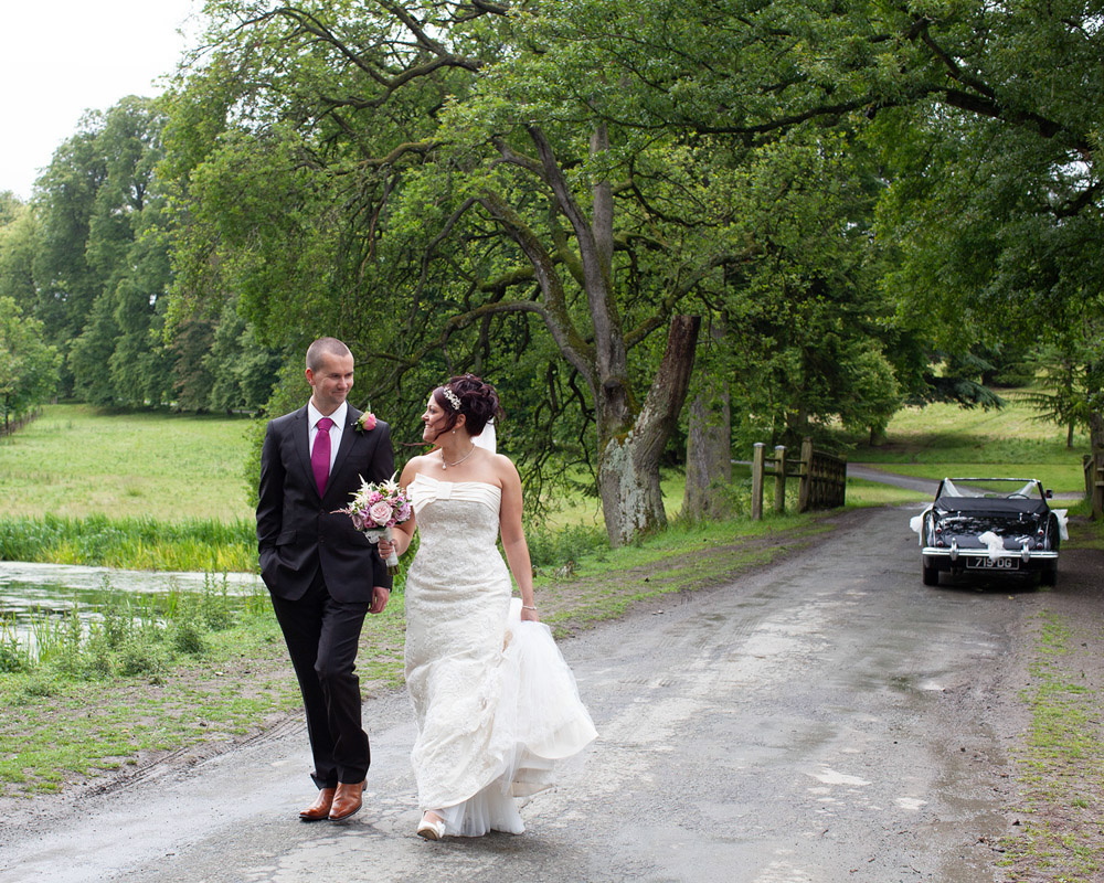Couple walk down a narrow road, smiling at each other with car in distance.