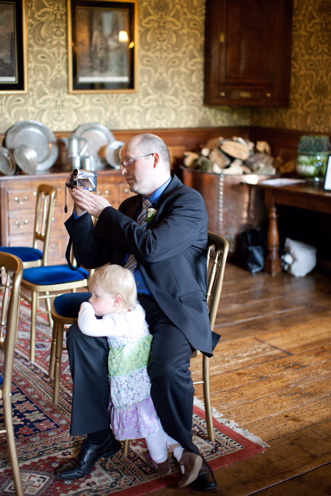 A man filming with a camcorder as his daughter leans gently on his knee