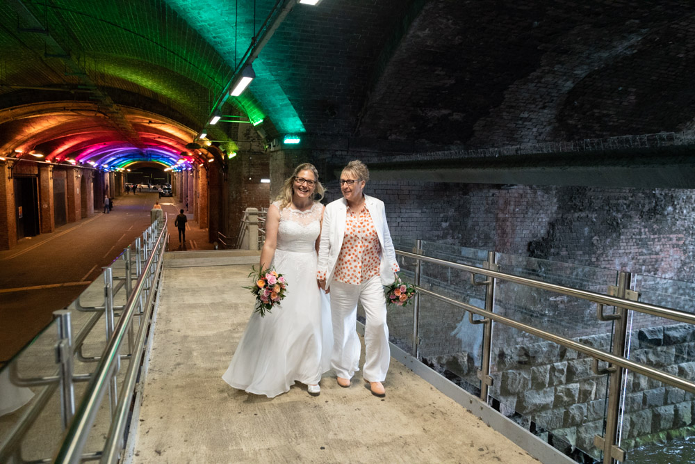 The couple continue along the Granary Wharf walkway with soft blue and pink lights behind