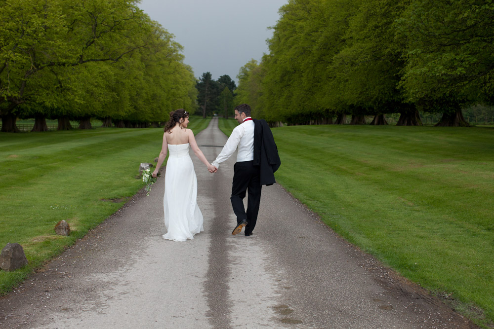 Bride and groom walking hand in hand, looking at each other along a narrow road lined with trees