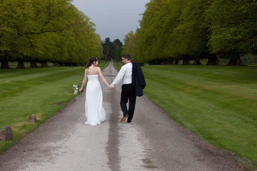 Bride and groom walking hand in hand, looking at each other along a narrow road lined with trees