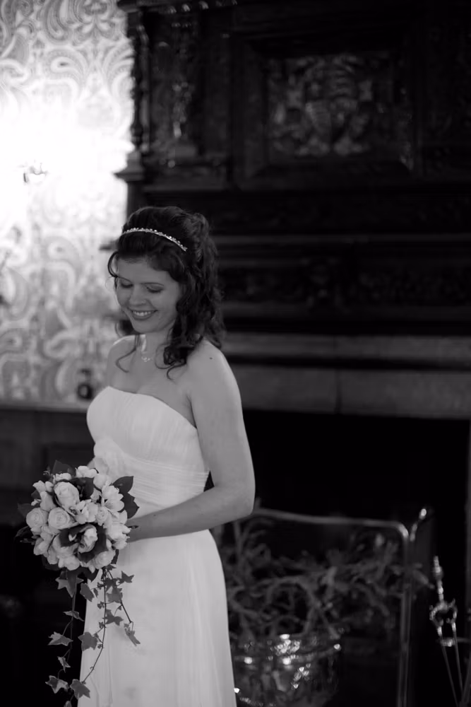 Bride standing by the fireplace indoors, softly looking down at her bouquet