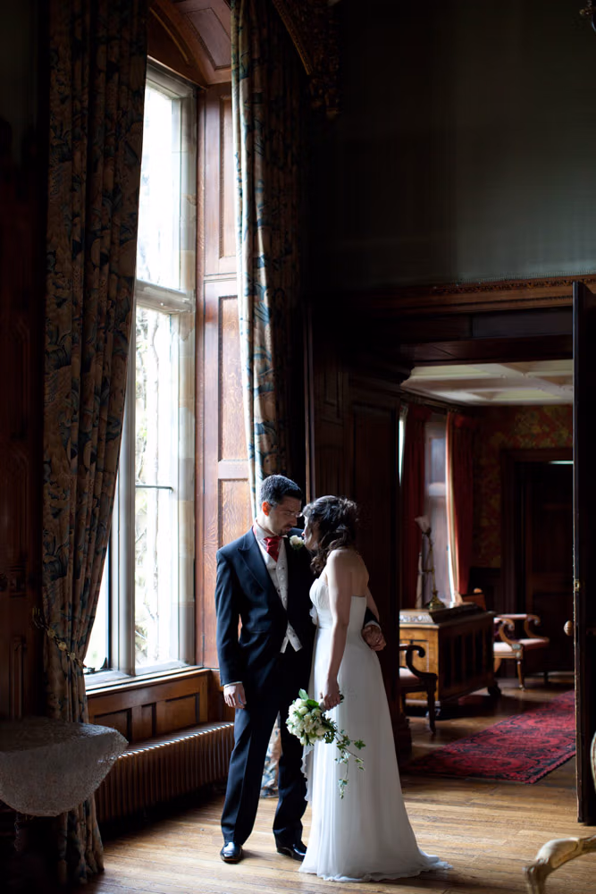 Bride and groom standing face to face beside a tall window with draping curtains
