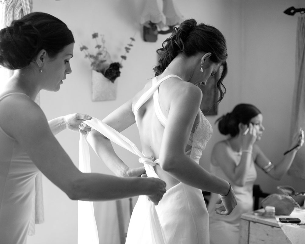 Bridal prep moment — one bridesmaid ties the dress while another does her makeup nearby.