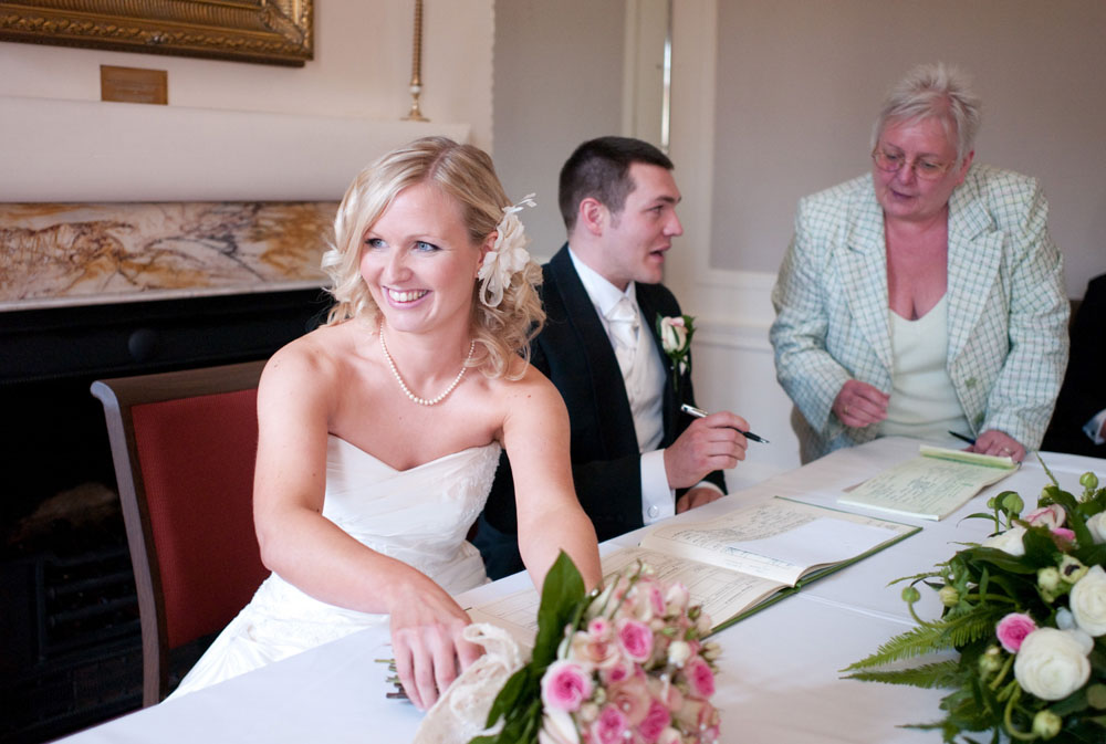 Bride smiling at her bridesmaids while the groom signs the register at Hoyle Court, West Yorkshire wedding