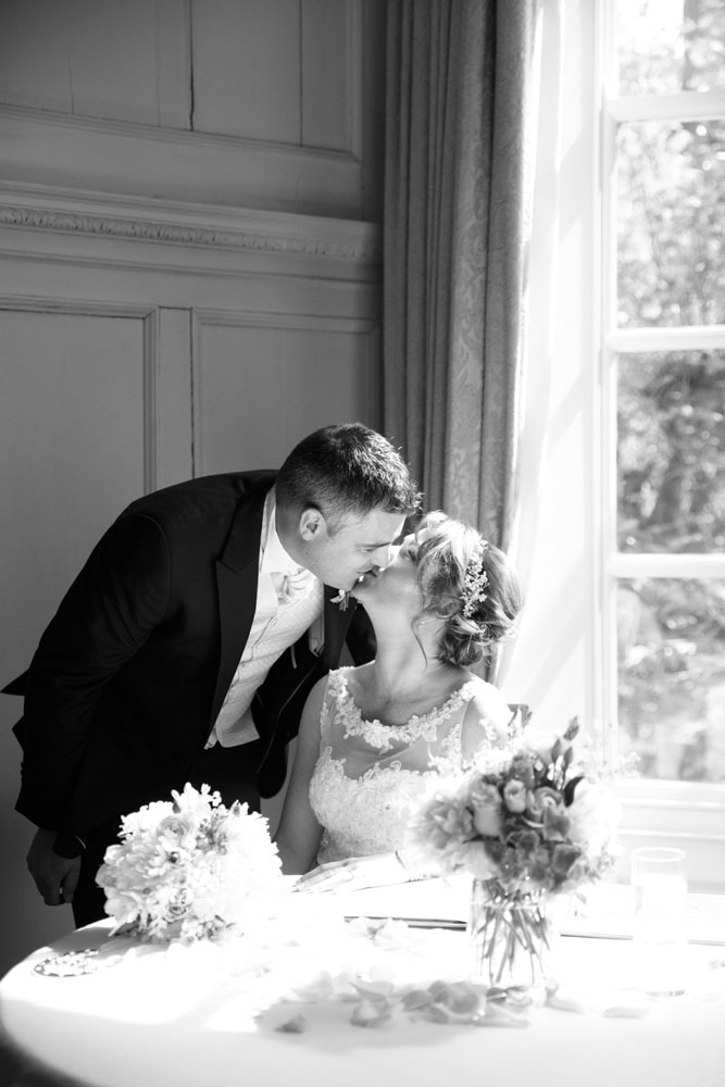 Newlyweds share a kiss in soft sunlight, captured in timeless black and white.